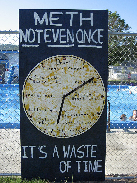 Sign made of  4’X8’ sheets of plywood.  Background spray painted in navy blue and brush painted with exterior enamel white paint for lettering.  Clock made out of sheet of plywood, 45 inches in diameter.  Clock was roller painted with exterior enamel white paint and the yellow exterior paint on the clock face was dabbed on with a rag.  The lettering on the clock was brush painted with black and red exterior enamel paint. Lettering on the clock face is symptoms of Meth use.  Hands on the clock are made from a metal yard stick, spray painted navy blue.  Clock is anchored to background with screws. Inspiration for the clock, was how little time passes before you are in trouble with meth, when you use it.